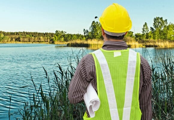 Engineer in safety gear inspecting a water body outdoors.