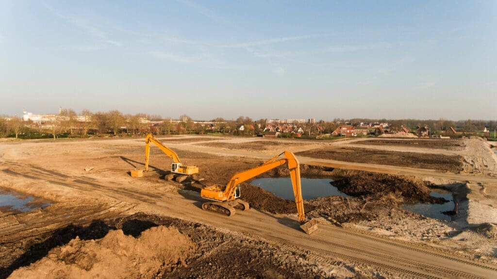 Two excavators digging in a large, dry construction site under a clear sky.