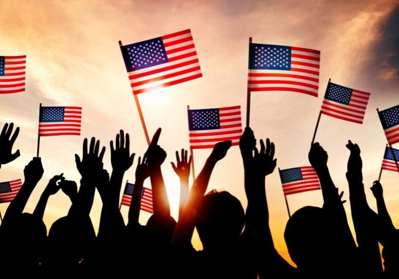 Silhouetted crowd waving American flags at sunset.