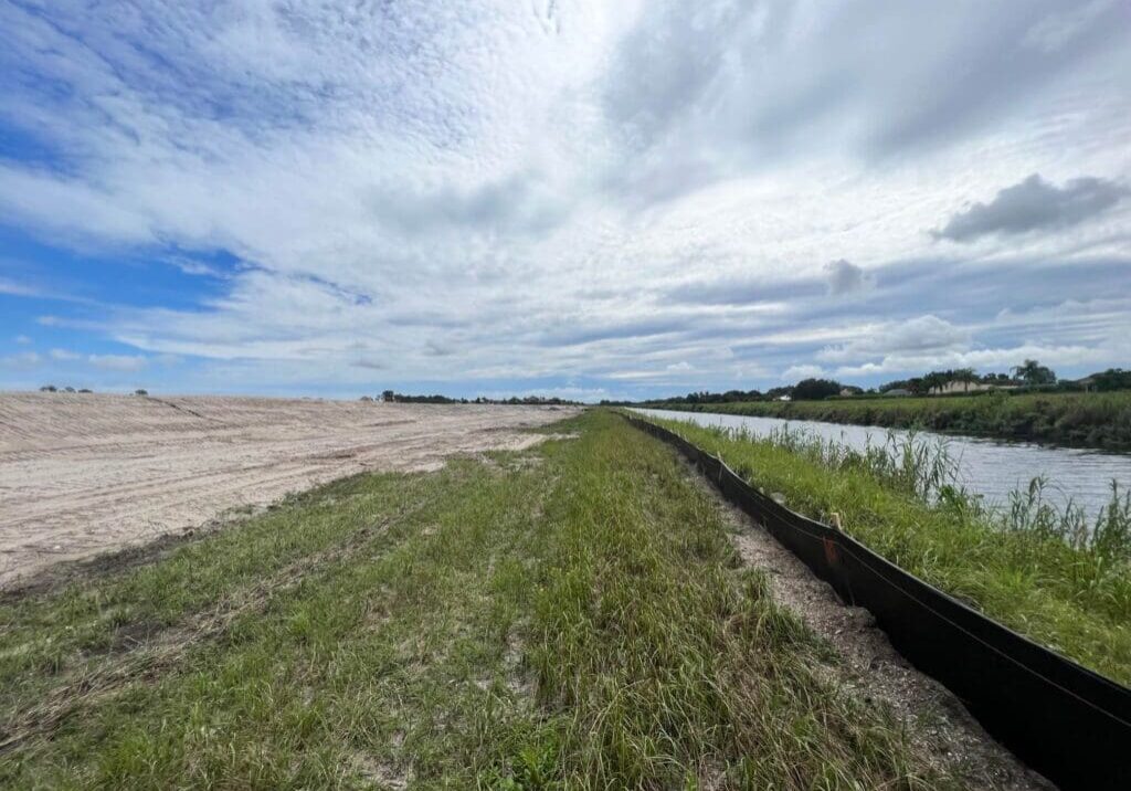 A grassy embankment between sandy land and a water canal under a cloudy sky.