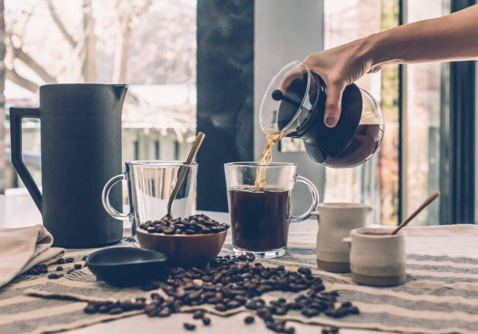 Pouring coffee into a glass mug surrounded by coffee beans and cups.