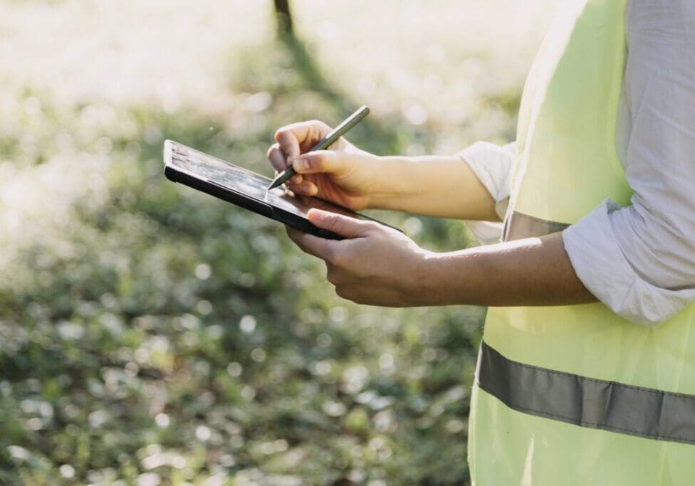 Person in safety vest writing on clipboard outdoors.