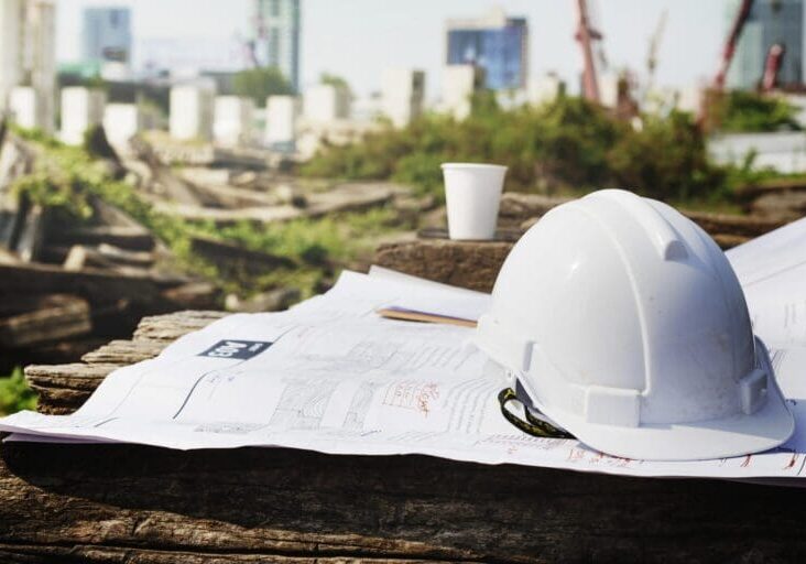 Construction plans and a white safety helmet on a wooden surface outdoors.