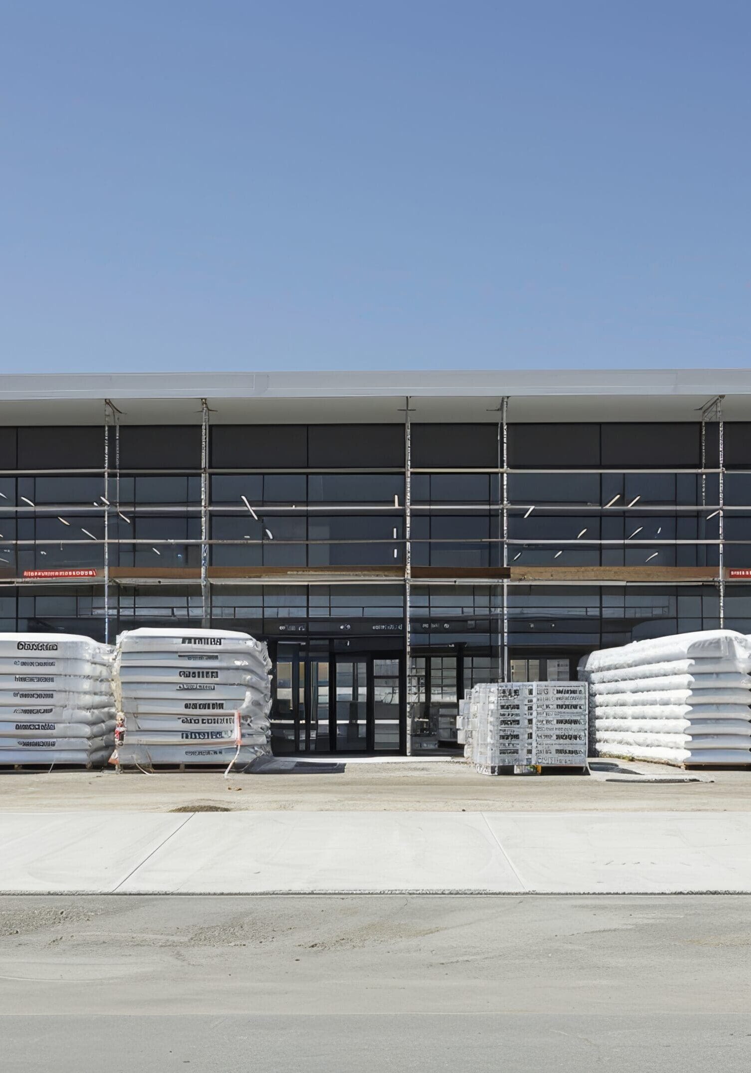 Stacks of packaged materials in front of a large industrial building under clear blue sky.