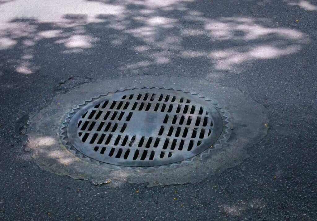 Round metal storm drain grate on asphalt road with shadows of tree leaves.