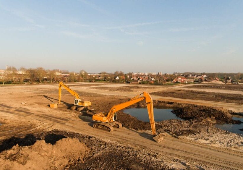 Two excavators digging in a large, dry construction site under a clear sky.