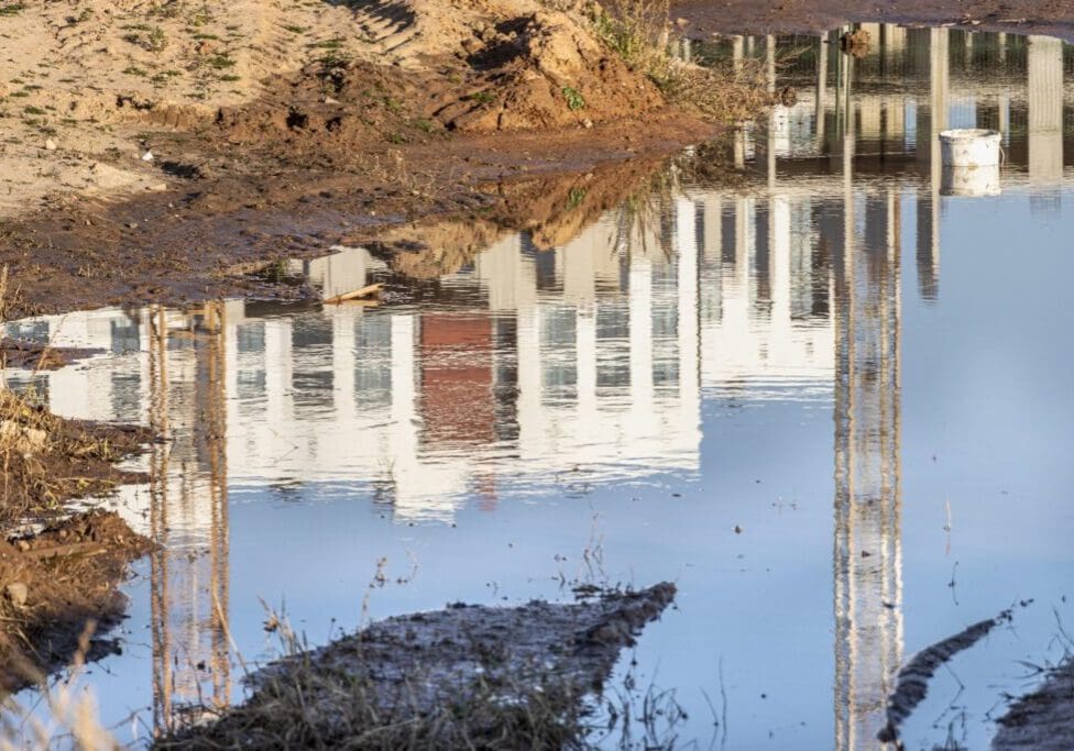 Reflection of a building in calm water with mud and grass around.