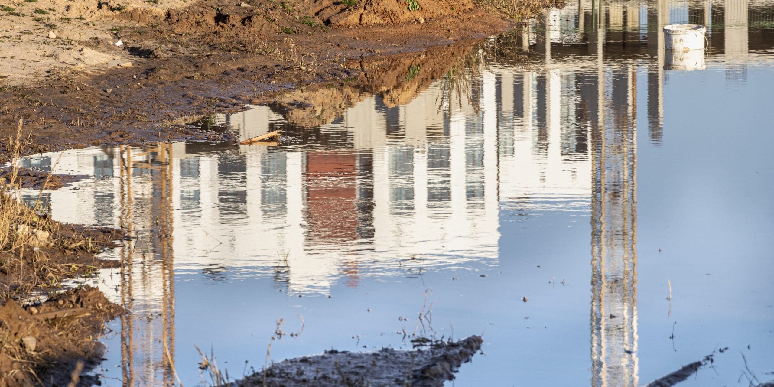 Reflection of a building in calm water with mud and grass around.