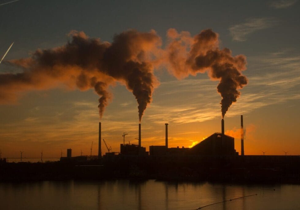 Industrial factory chimneys emitting smoke at sunset over a body of water.