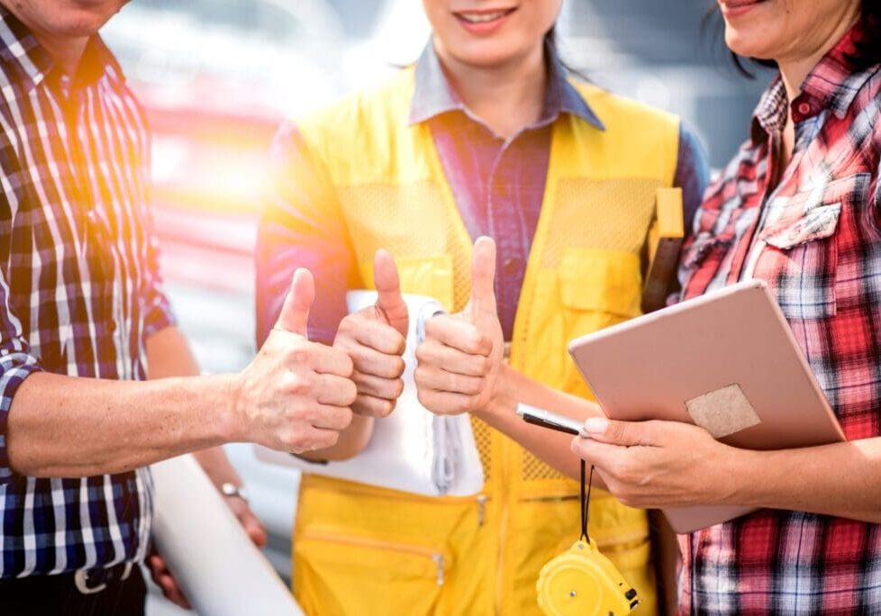 Group of workers in safety gear giving thumbs up in a bright industrial setting.