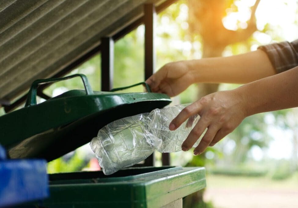 Person recycling plastic bottle by placing it in a green bin outdoors.