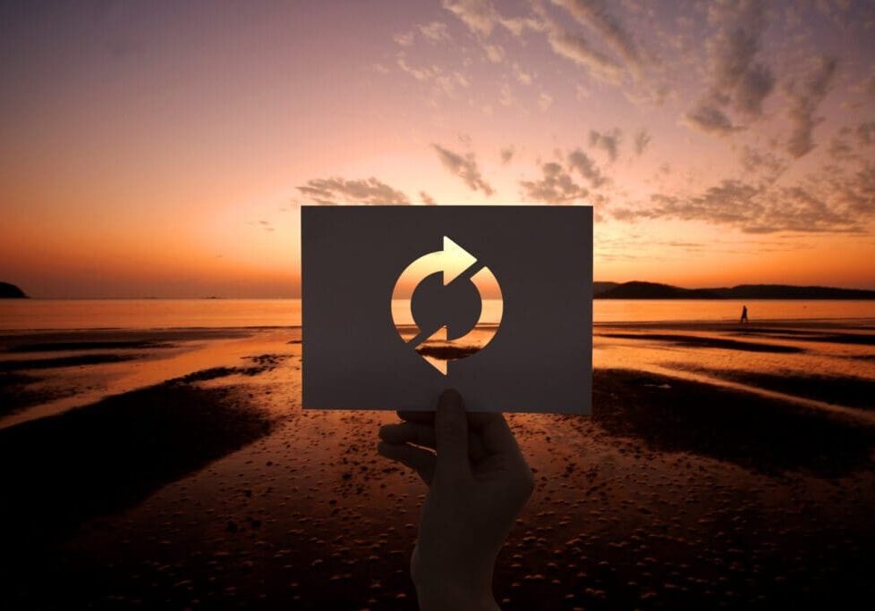 Hand holding a card with a recycle symbol against a sunset at the beach.