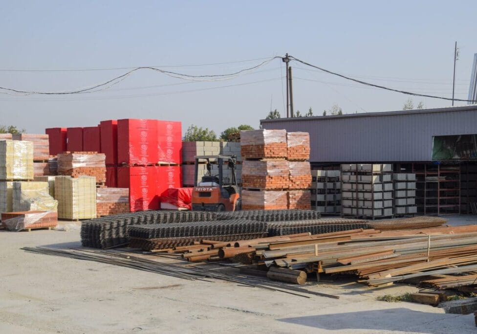 Construction materials like bricks, steel rods, and red containers are stacked outdoors.