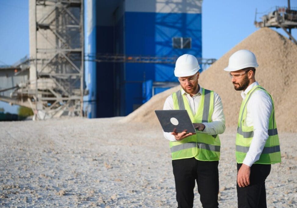 Two construction workers reviewing plans on a laptop at a site.