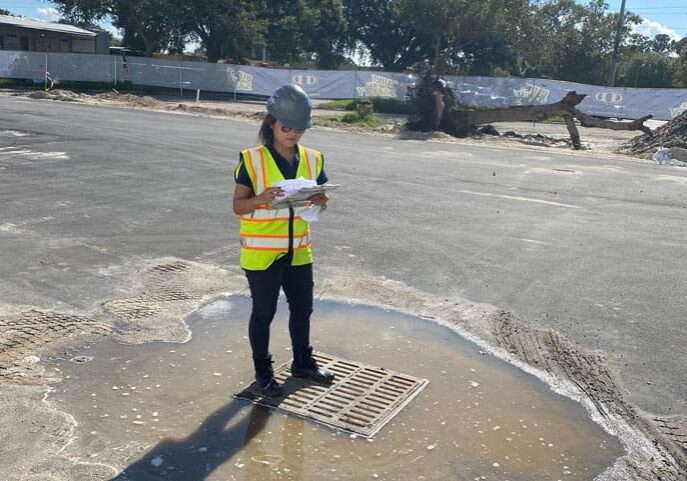 A person in a yellow safety vest and helmet inspecting a site with a clipboard.